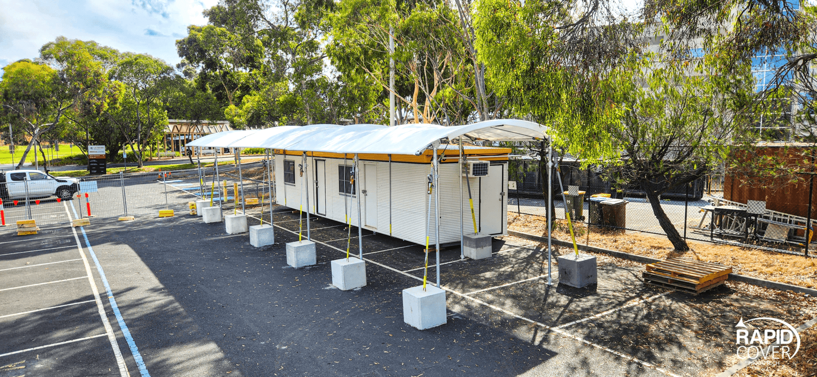 A white portable building with a curved canopy roof stands in a parking lot, supported by yellow poles and concrete blocks, surrounded by trees and a few benches; "Rapid Cover" logo in the corner.