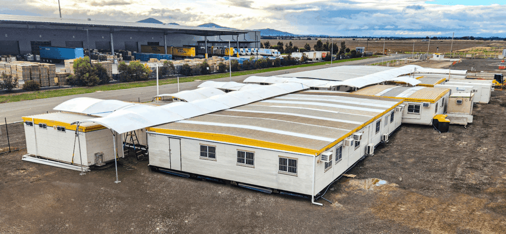 A logistics area with storage containers featuring white roofs, a ladder, and various pallets stacked nearby. Trees are visible in the background.