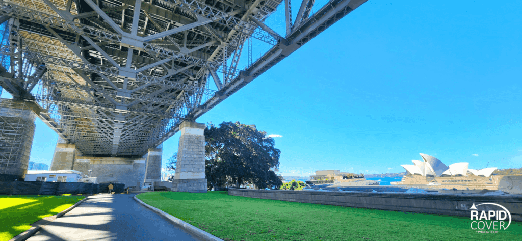 View from beneath the Sydney Harbour Bridge, showing its steel structure above a walkway and green lawn, with the Sydney Opera House and blue sky visible in the background. Rapid Cover logo in the bottom right corner.