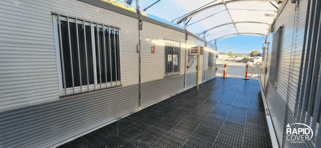 A covered walkway with metal flooring connects portable modular buildings with barred windows. The area is clean and well-lit, and the Rapid Cover logo is visible in the bottom right corner.