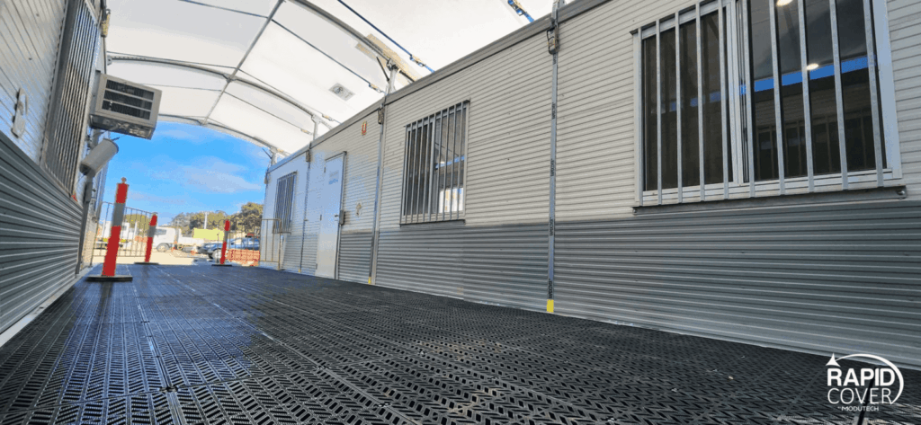 A covered outdoor walkway with grated flooring leads past portable buildings with barred windows and white doors. The Rapid Cover logo is visible in the lower right corner. Bright blue sky is visible outside.