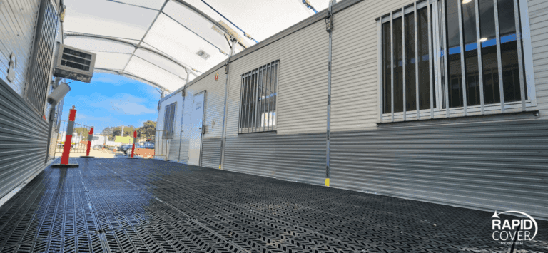 A covered outdoor walkway with grated flooring leads past portable buildings with barred windows and white doors. The Rapid Cover logo is visible in the lower right corner. Bright blue sky is visible outside.