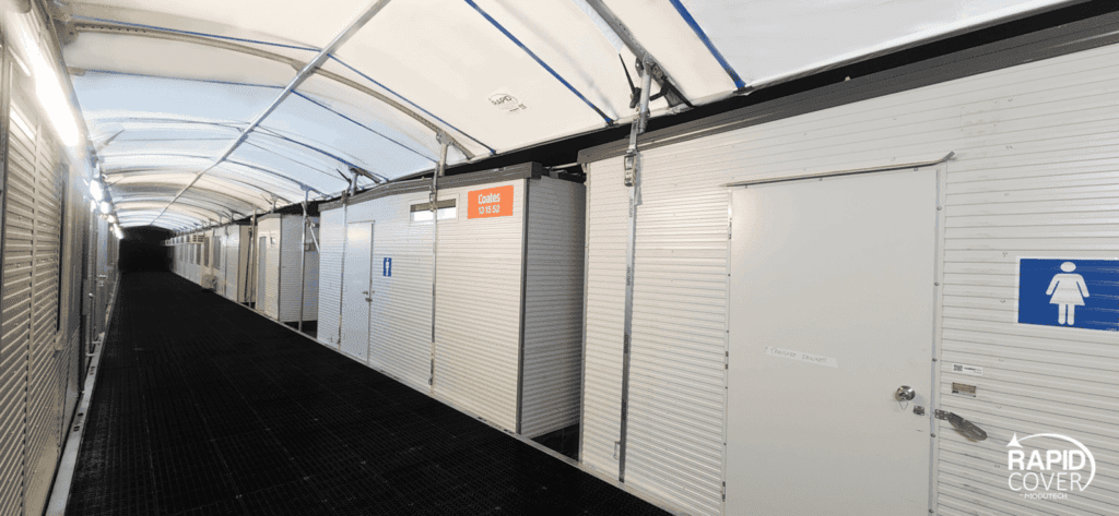 A brightly lit corridor with white, portable restroom units on the side, including a women’s restroom marked by a blue sign. The ceiling is covered with a translucent, arched canopy. "Rapid Cover" logo is visible.