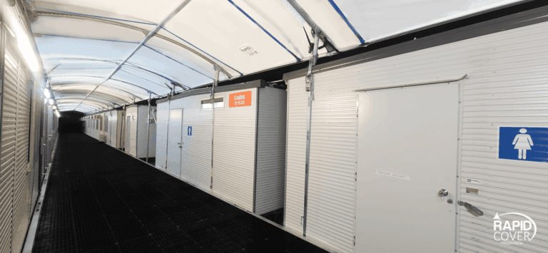 A brightly lit corridor with white, portable restroom units on the side, including a women’s restroom marked by a blue sign. The ceiling is covered with a translucent, arched canopy. "Rapid Cover" logo is visible.