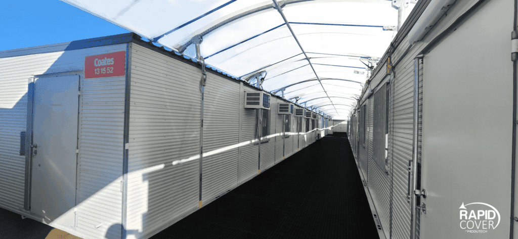 A covered outdoor hallway with portable cabins on both sides, each having doors and windows. Sunlight shines through the white translucent roof. The Rapid Cover logo appears in the lower right corner.