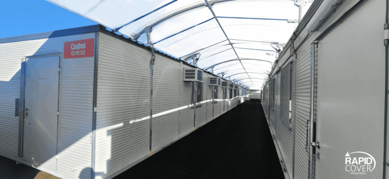 A covered outdoor hallway with portable cabins on both sides, each having doors and windows. Sunlight shines through the white translucent roof. The Rapid Cover logo appears in the lower right corner.