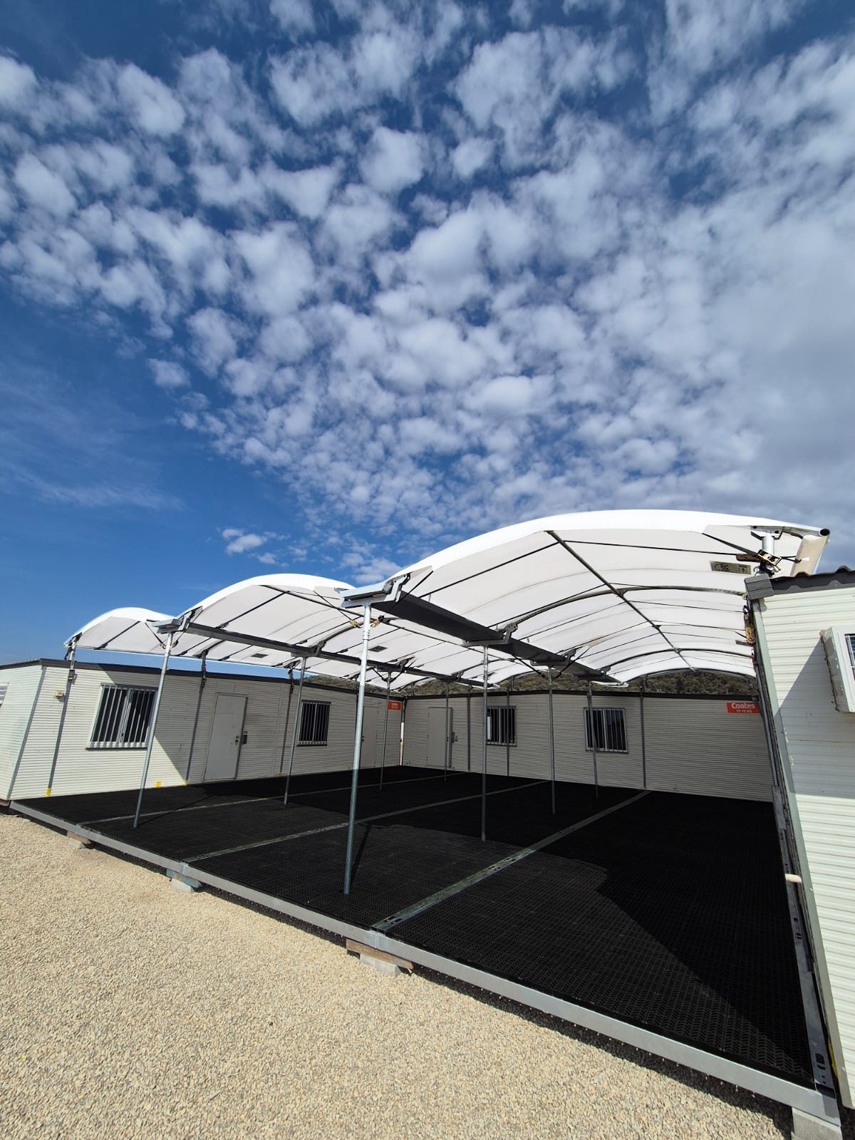 A large outdoor structure with a white, curved canopy roof and open sides stands on a gravel surface under a blue sky with scattered clouds. The design is ideal for sustainable temporary walkway covers, bordered by white buildings with barred windows.