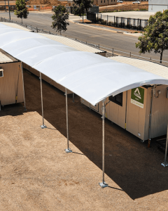 A white, curved canopy is installed over a walkway beside a long, portable building on a sunny day. The structure is supported by metal poles, with a tree and road in the background.