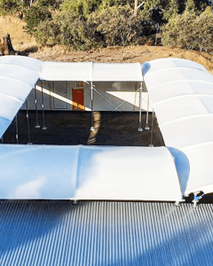 Aerial view of a square structure with a central open courtyard, featuring white curved fabric roofs and metal roofing, surrounded by dry grass, trees, and paved areas.
