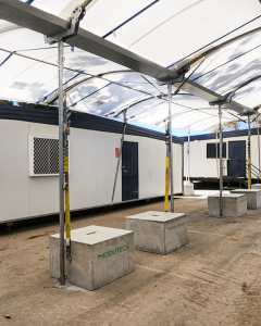 A temporary outdoor structure with a white canopy covers a walkway. In the background, there's a portable building with windows and a door. The area is supported by metal poles and surrounded by rectangular concrete blocks.