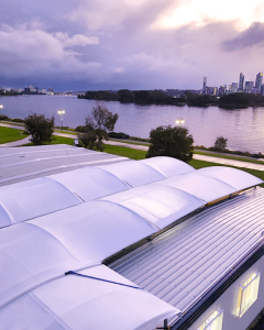 View of a river with a city skyline in the background, taken from a rooftop with curved solar panels at sunset. The sky is partly cloudy, and the water reflects the soft hues of dusk.