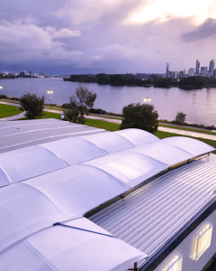 A large white-roofed modular building sits by a river, with a green park on one side and a city skyline under a dramatic cloudy sky at sunset. The "Rapid Cover Productions" logo appears in the lower right corner.