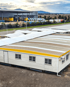 A logistics area with storage containers featuring white roofs, a ladder, and various pallets stacked nearby. Trees are visible in the background.