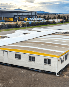 A row of portable office buildings with yellow roofs and white shade canopies, set up on a dirt lot near an industrial warehouse and surrounded by construction materials. The Rapid Cover Projects logo appears in the lower right corner.