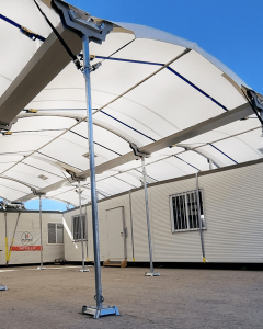A large white canopy with metal supports is installed over a portable building against a clear blue sky. The building has small windows with bars and a door visible beneath the canopy.