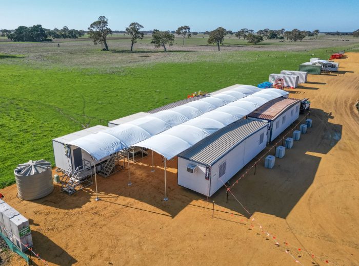 A row of temporary white structures and portable buildings on a dirt area, surrounded by green grass and scattered trees, with blue skies overhead. Sustainable temporary walkway covers, barriers, and water tanks are visible nearby.